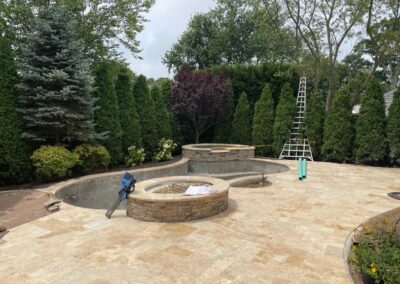 A backyard landscaping project underway showcases a partially built in-ground pool paired with a nearby stone hot tub. Amidst ladders and construction tools, the area is encircled by tall trees and newly laid stone tiles, setting the stage for a serene outdoor retreat.