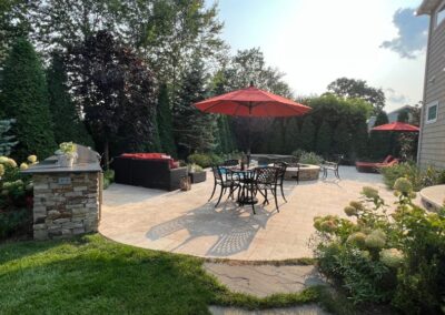 A spacious backyard patio features a central table and chairs under a red umbrella. Nearby, a stone firepit with seating invites relaxation. Expert back yard landscaping with lush greenery and tall trees surrounds the area, crafting a private and tranquil atmosphere.