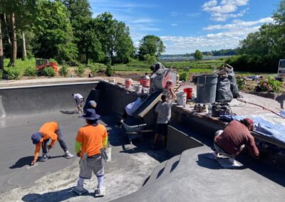 Workers in bright shirts and hats apply concrete to the interior of an empty pool, transforming it into a back yard landscaping masterpiece. Various tools and buckets are scattered around. The background features green trees, a blue sky, and a view of water in the distance.