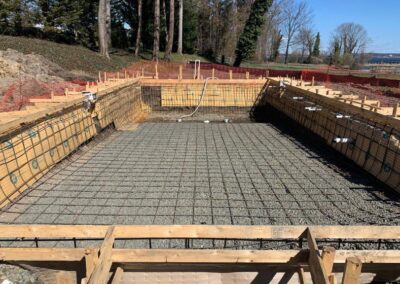 The construction site, resembling a back yard landscaping project, showcases an early-stage concrete foundation with reinforcement bars in a rectangular pit. Wooden formwork surrounds the area, while trees stand tall in the background under a clear blue sky.