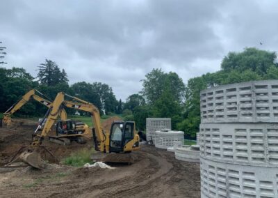A construction site features two yellow excavators working on soil, reminiscent of back yard landscaping. Nearby are large concrete drainage pipes, hinting at functional yet aesthetic outdoor transformations. The background shows lush green trees under a cloudy sky.
