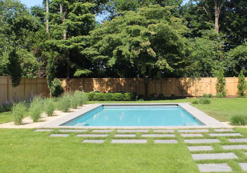 A rectangular backyard swimming pool is surrounded by green grass, stepping stones, and landscaping with bushes and trees, enclosed by a wooden fence.