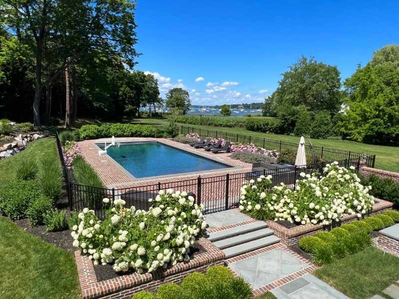 A rectangular swimming pool surrounded by brick paving, black fencing, blooming white flowers, lounge chairs, and lush green lawns, with trees and a lake visible in the background under a blue sky.