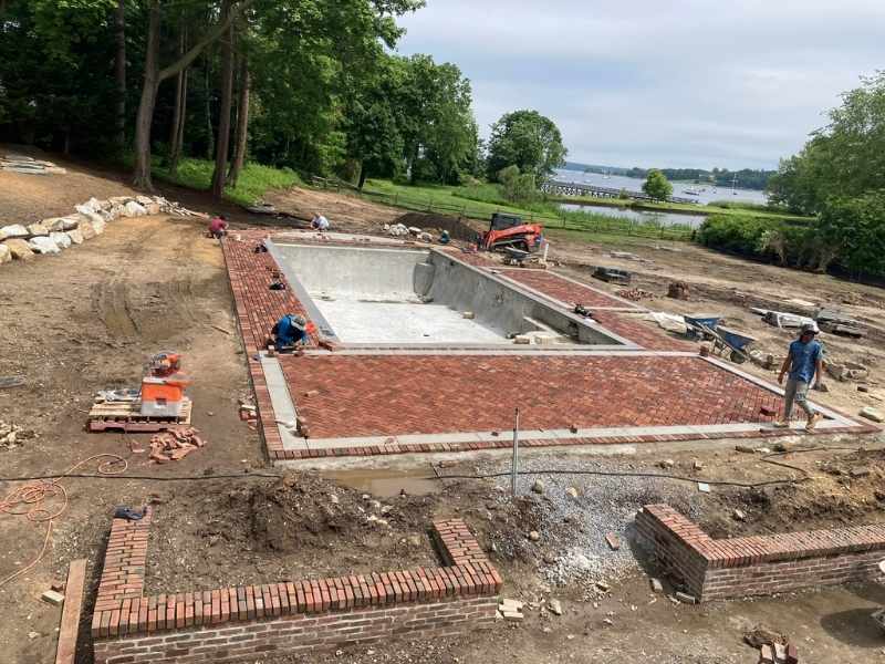 Workers are constructing a rectangular brick patio around an in-ground pool on a large property with trees and a distant body of water in the background. Construction equipment and bricks are scattered around the site.