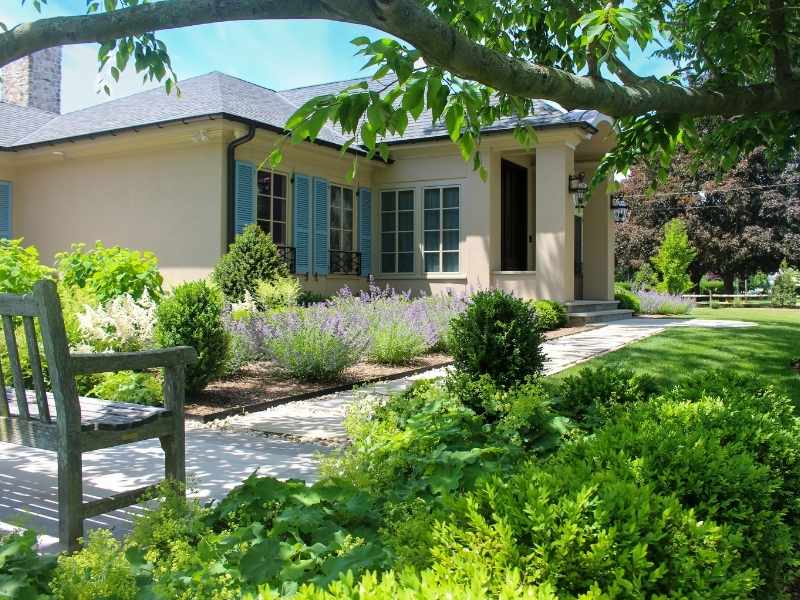 A single-story house with light-colored walls and blue shutters is surrounded by a lush, green garden with shrubs, flowering plants, a stone pathway, and a wooden bench under a tree.