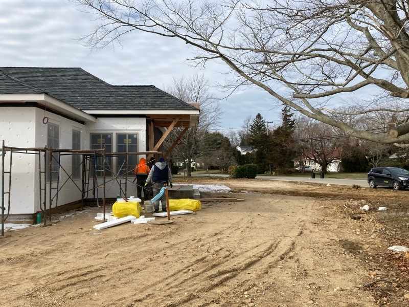 Two workers stand on scaffolding and work on the exterior of a house under construction. Yellow insulation material and tools are on the ground beside them; the yard is bare dirt with tire tracks and scattered debris.