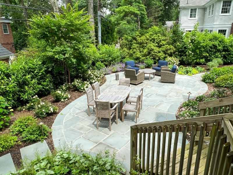 A backyard patio with a stone floor features a dining table with six chairs and a separate seating area with blue cushioned chairs, surrounded by lush greenery and flowering plants. Wooden steps lead down to the patio.