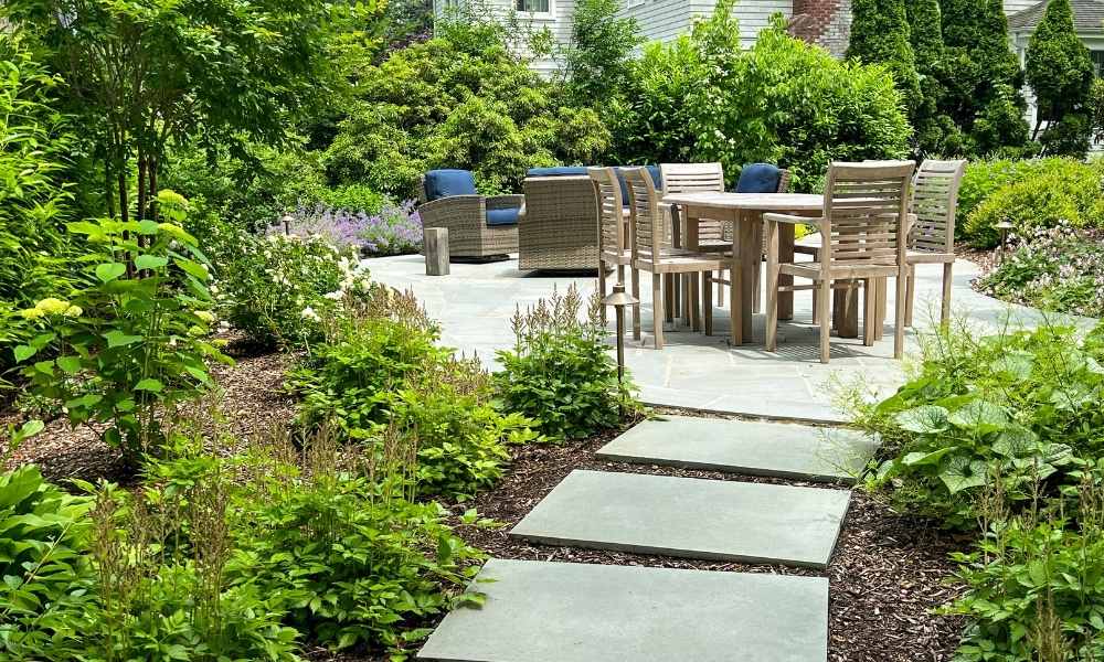 Stone path leads to a patio with a wooden dining table and chairs, surrounded by lush greenery and shrubs. Outdoor lounge chairs with blue cushions are also visible on the patio.